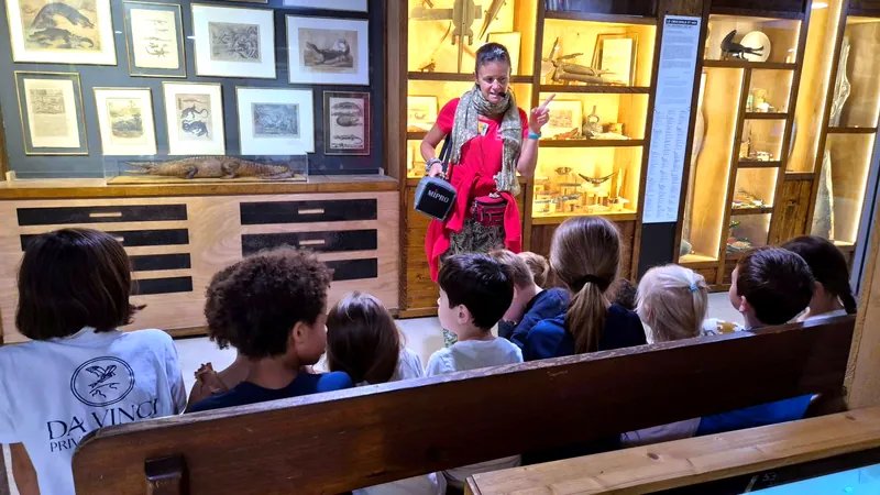 Assis sur un banc, les enfants écoutent la guide de la Ferme aux Crocodiles devant des vitrines de gravures et d'objets sur les crocodiles