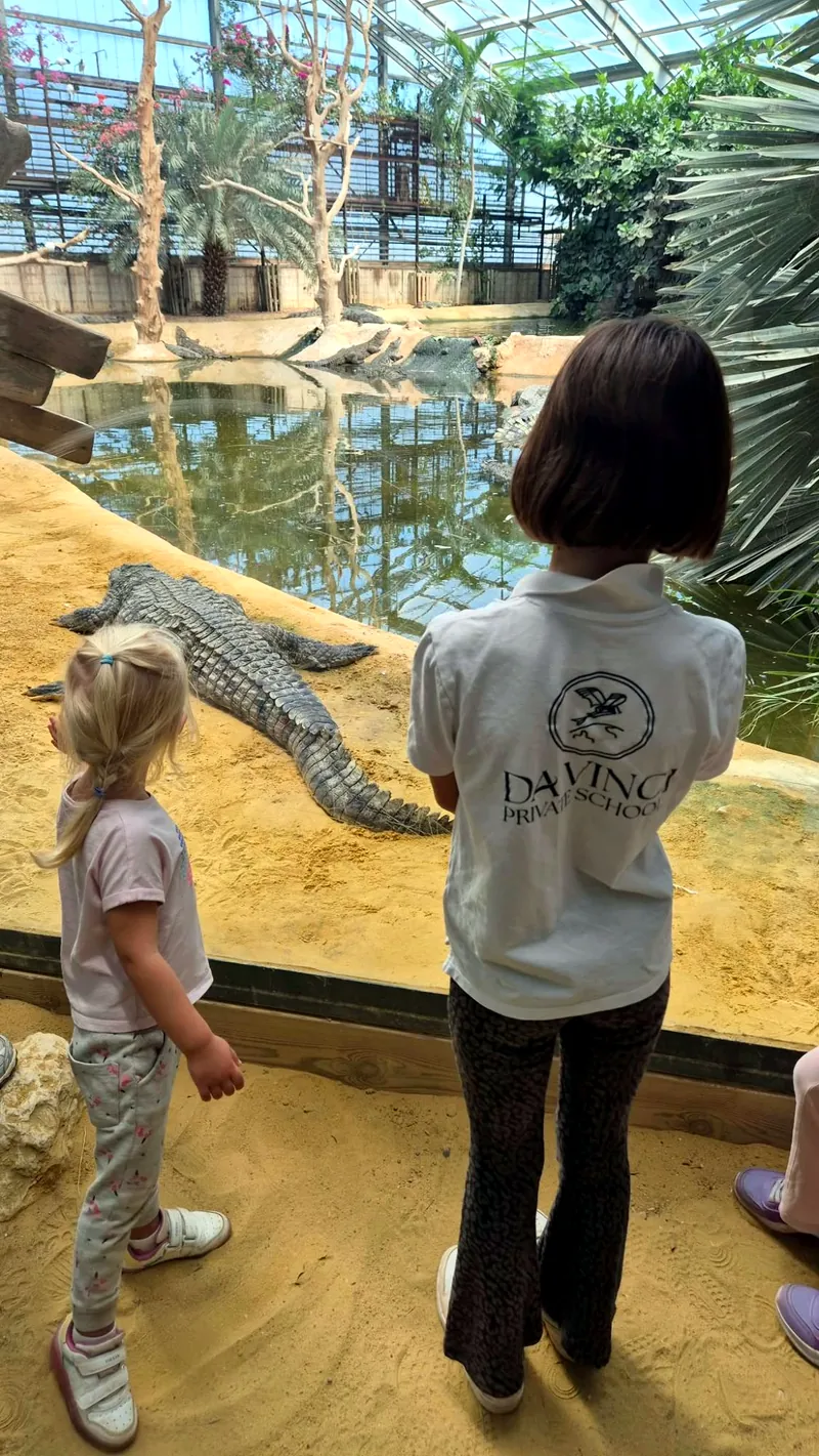 Deux fillettes de l'école, de dos, regardent un crocodile du Nil allongé sur le sable au bord du bassin