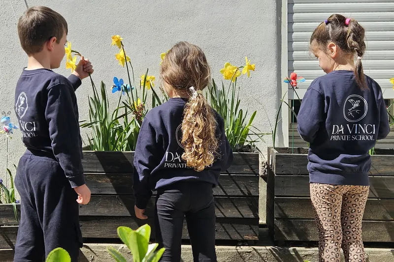 Trois enfants en sweat-shirt Da Vinci Private School, de dos, devant les jardinières de jonquilles de l'école