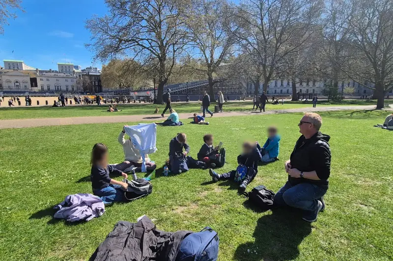 A group of children picnicking on the grass of a London park, with a teacher
