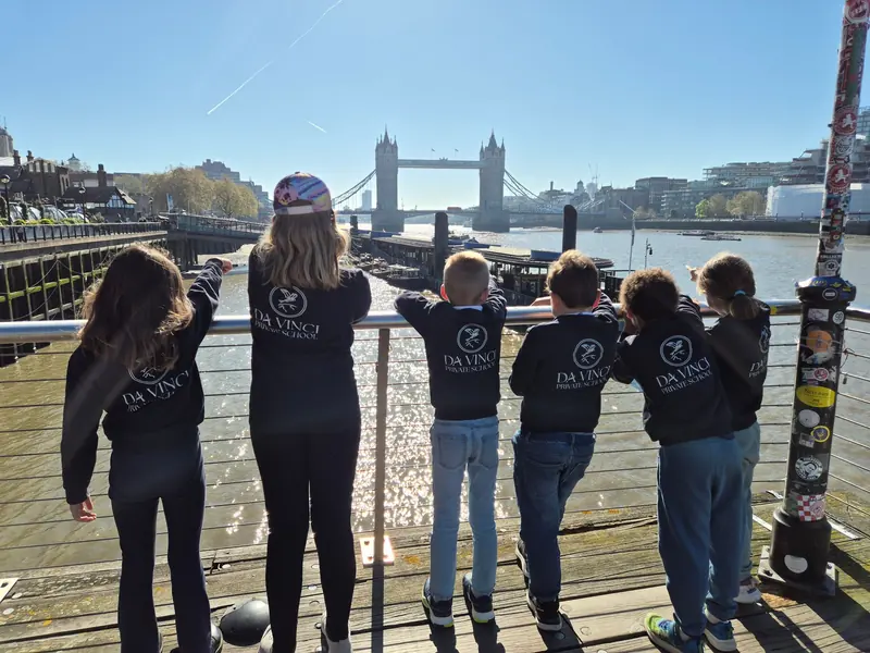 Six children in Da Vinci Private School sweatshirts, seen from behind, looking at Tower Bridge from a pontoon on the Thames
