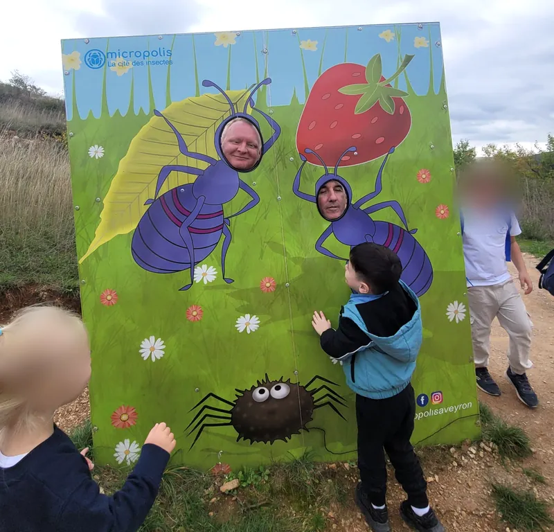 Deux enseignants passent la tête dans un panneau de fourmis de Micropolis, devant les enfants, sur un chemin en plein air