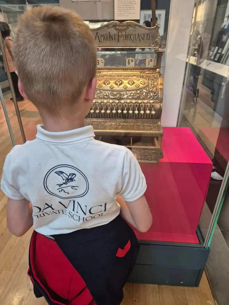 Un enfant en polo Da Vinci, de dos, observe une ancienne caisse enregistreuse dans un musée de Londres