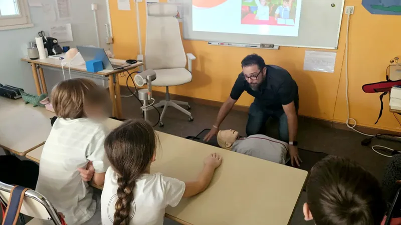 The trainer, kneeling by a first aid manikin, demonstrating a technique to children seated around the table